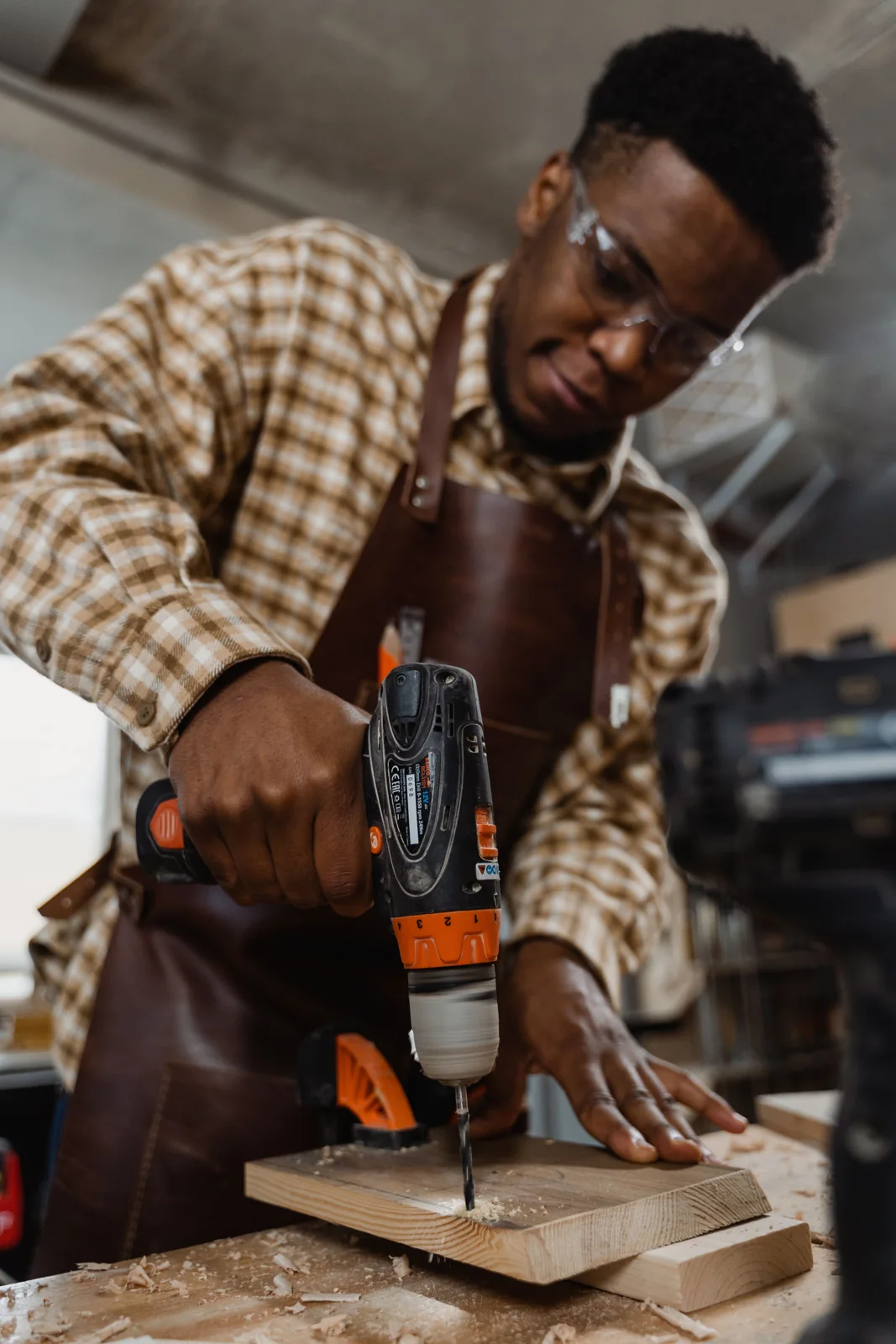 A woodworker wearing safety glasses drills into a clamped wood board while supporting it with one hand.