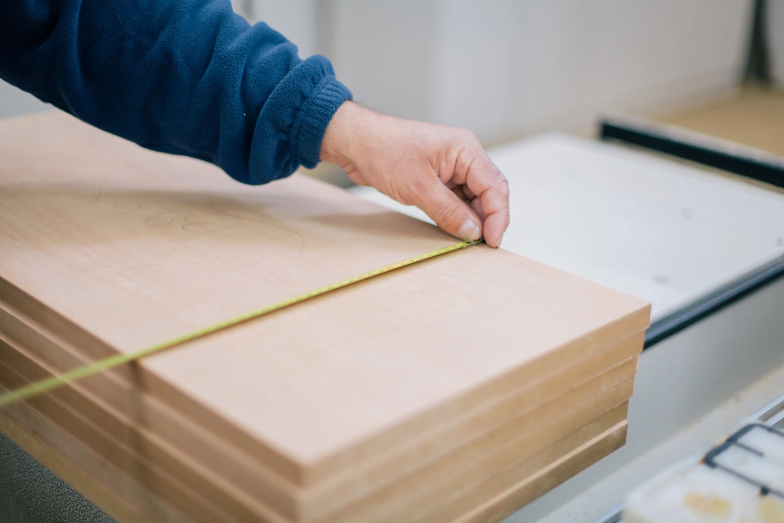 An adult hand stretches a yellow tape measure over the edge of a flat wood panel on a work surface.