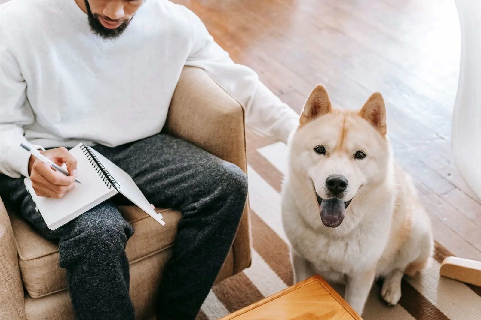 Man in an armchair writing notes in a spiral notepad while resting a hand on a happy Akita sitting beside him